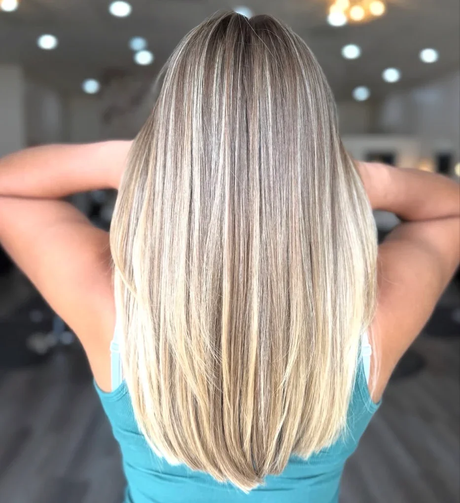 Back view of a woman with straight, shoulder-length blonde hair sitting in a salon with her arms raised.