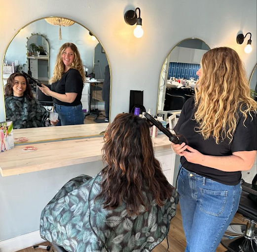 Hairstylist blow drying a woman's hair at a salon, with her smiling in a mirror.