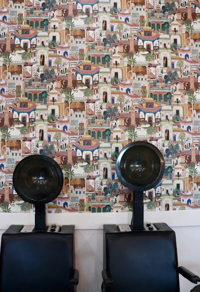 Two black salon hood dryers in front of a colorful, patterned wallpaper depicting a cityscape with buildings, trees, and arches.
