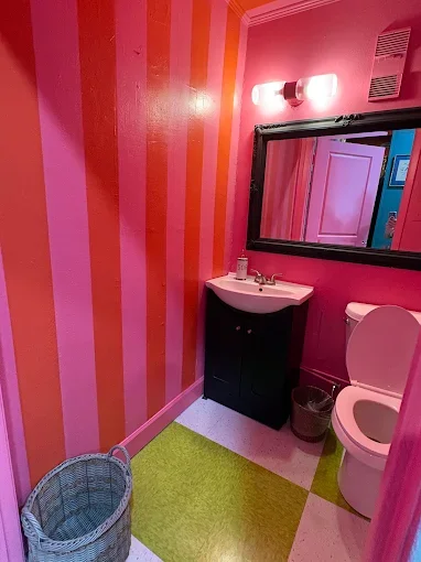 Small bathroom with pink and orange striped walls, black vanity with a sink, mirror with light above, toilet, green and white checkered floor, and a wicker basket.