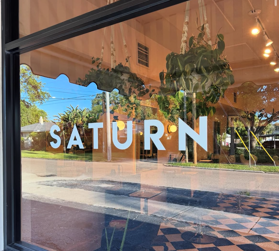 View through restaurant window with the word 'SATURN' in white letters, reflected street scene, green trees, and interior with hanging plant and checkered floor