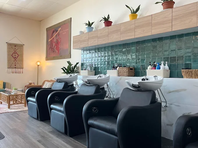 Salon with three black hair washing chairs in front of a green tiled wall, shelves with potted plants and hair products, and a sitting area with a rug and furniture.