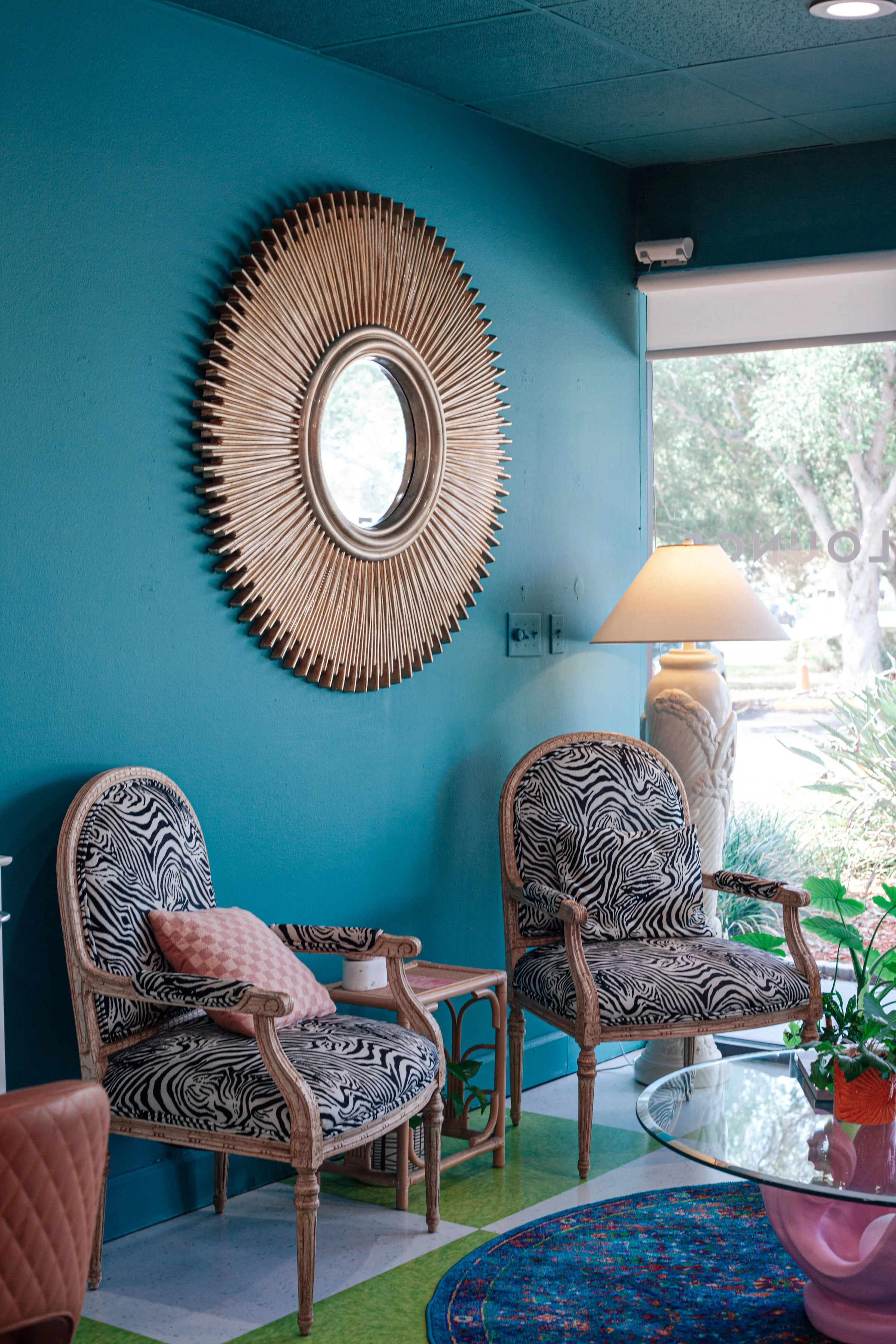 Interior of a room with teal walls, featuring two vintage-style chairs with black and white zebra print upholstery, a glass coffee table with a pink base, a tall white lamp, and decorative elements including a sunburst mirror on the wall and a window with a view of greenery outside.