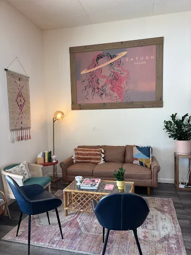 Interior of a salon with a pink and brown color scheme, featuring a pink sofa, two blue chairs, a wooden coffee table, a wall hanging, a floor lamp, a large framed artwork of a woman with pink hair and a planet ring, and a potted plant.