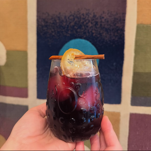 Hand holding a glass of red wine with ice, garnished with a lemon slice and cinnamon stick, with a colorful abstract background.