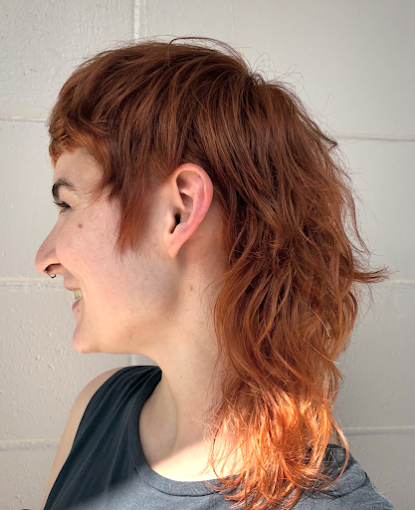 Profile of a smiling woman with curly, shoulder-length red hair, wearing a sleeveless shirt, standing against a light-colored brick wall.