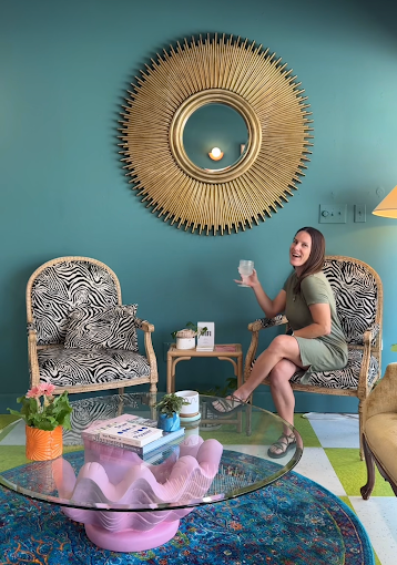 A woman sitting on a zebra-print armchair in a colorful living room, holding a drink and smiling. The room has a large decorative sunburst mirror on a teal wall, a glass coffee table with pink decorative objects, a small side table with plants, and a