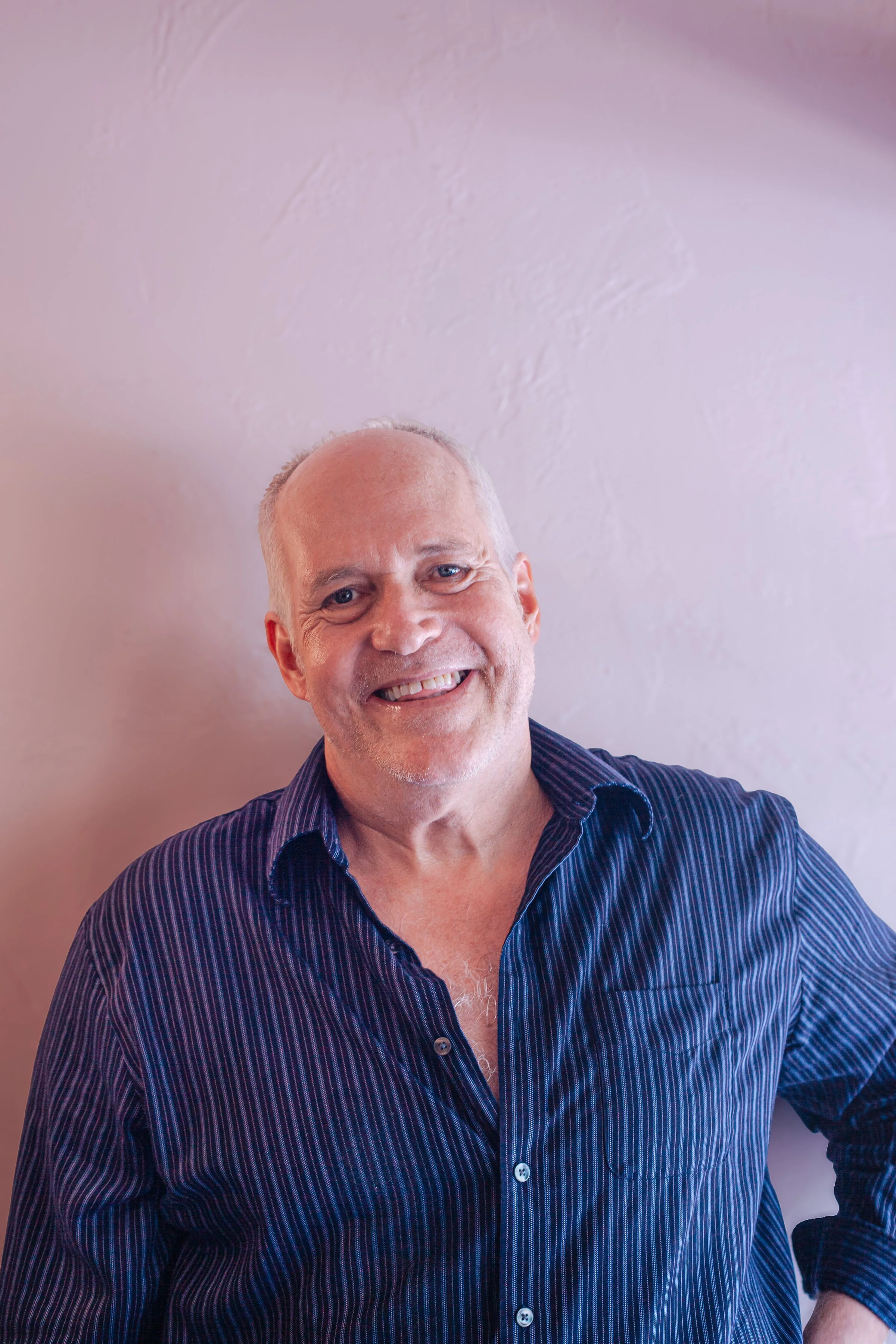Close-up of a smiling bald man wearing a dark striped shirt, standing against a light-colored textured wall.