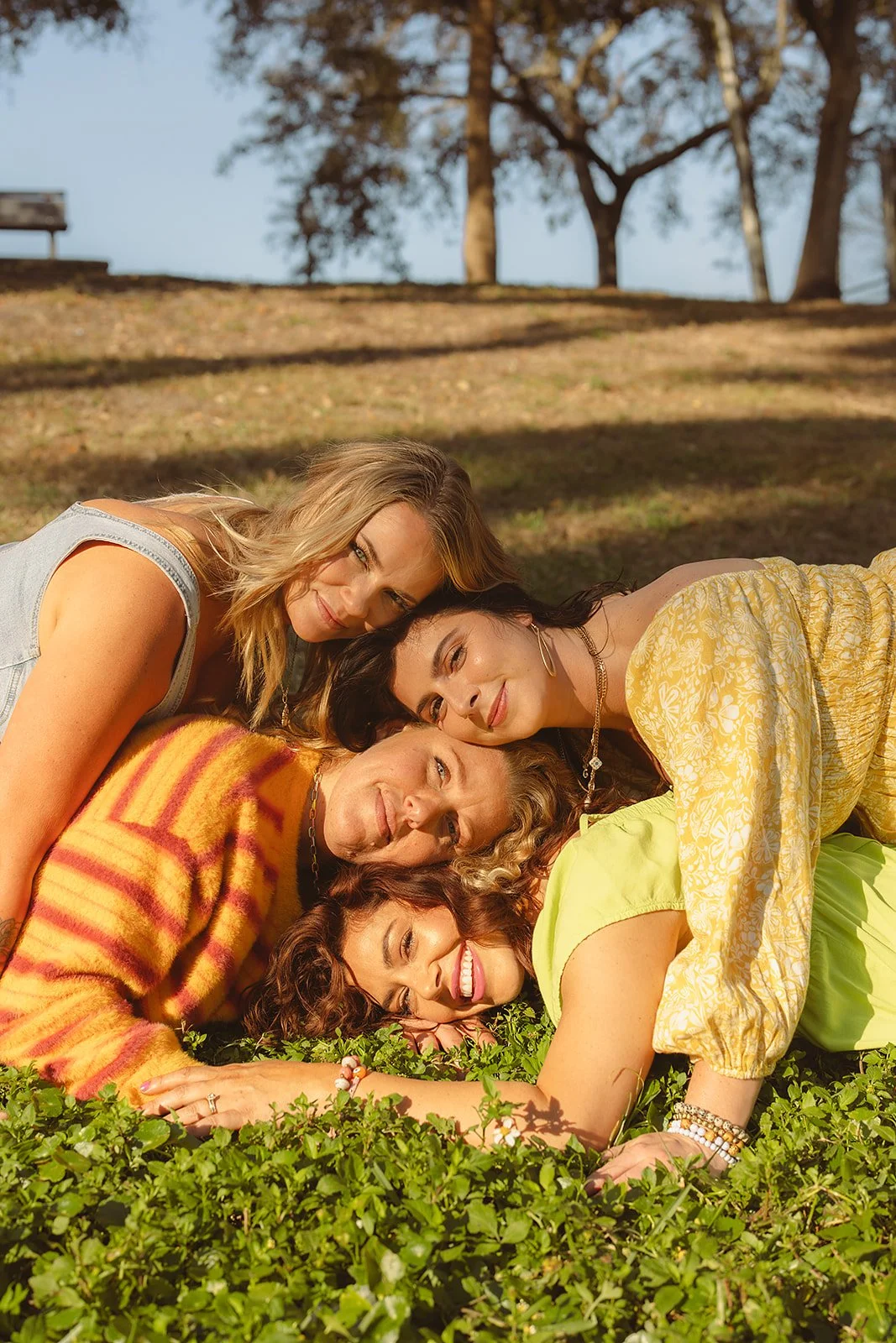 Four friends lying on the grass in a park, smiling and cuddling together during sunset.