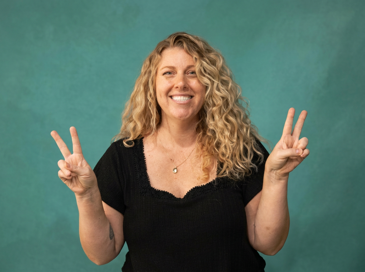 A woman with blonde curly hair smiling and holding up peace signs with both hands against a green background.