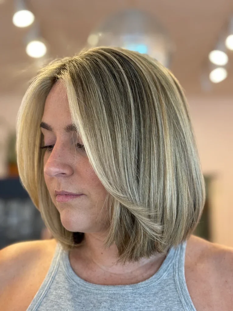 A woman with shoulder-length blonde hair styled in a sleek bob, wearing a gray sleeveless top, in a salon or similar setting.