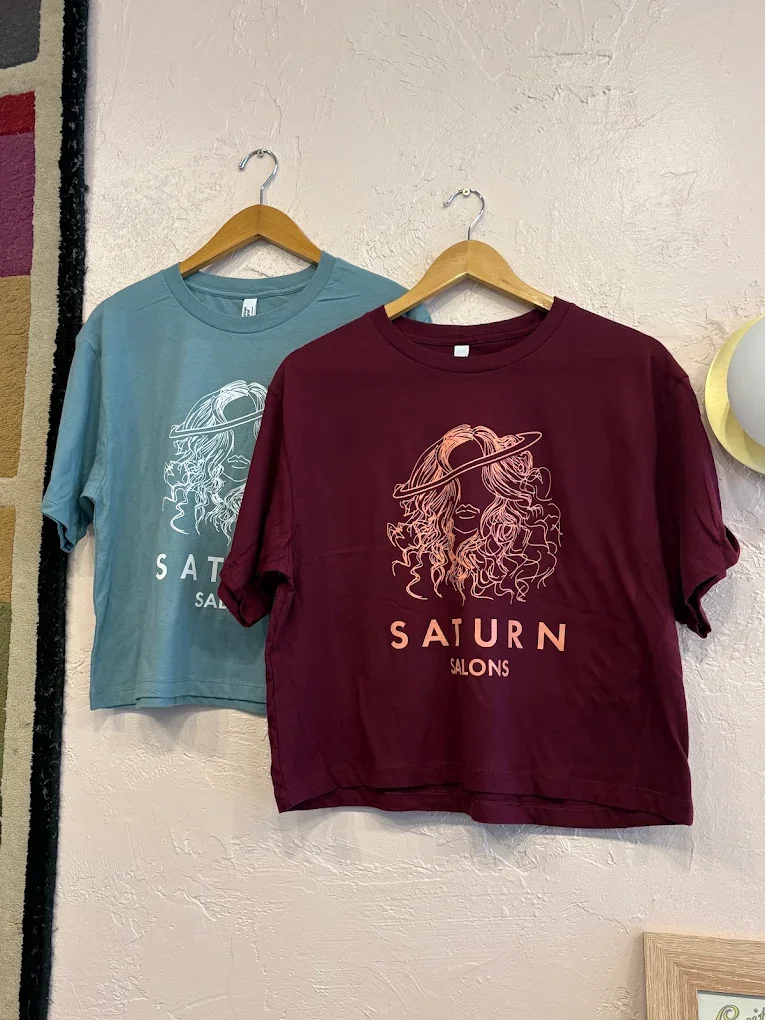 Two T-shirts hanging on wooden hangers on a white textured wall. The T-shirts are from Saturn Salons; one is light blue with a white line drawing of a woman's face with curly hair and a headband, and the other is maroon with a similar line drawing of
