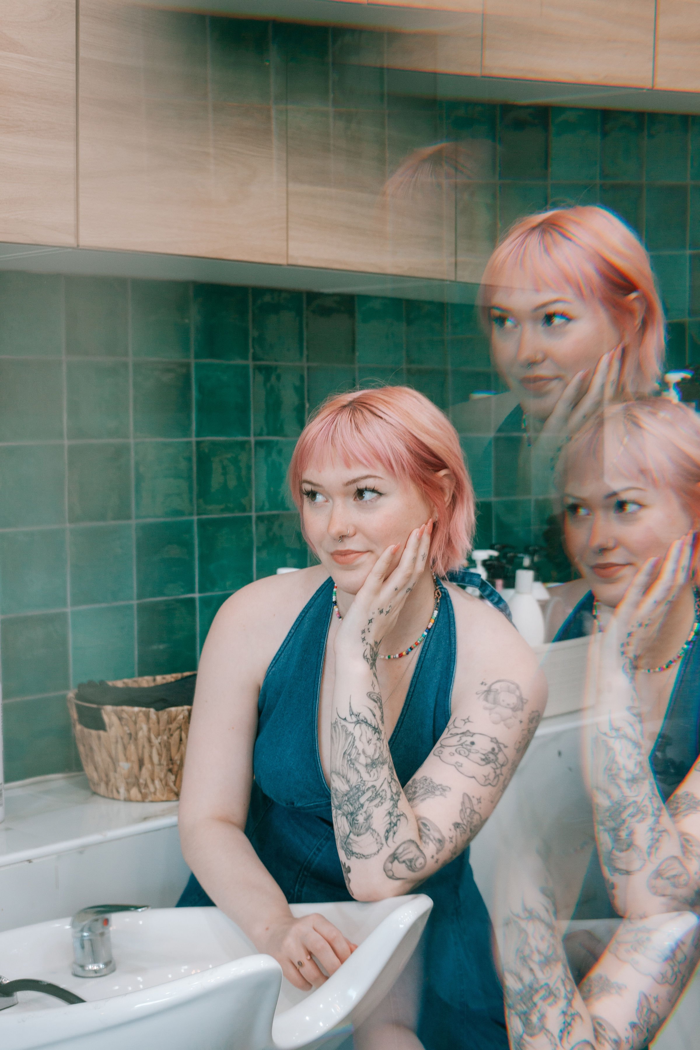 A young woman with pink hair, tattoos on her arm and chest, wearing a blue sleeveless top, standing at a bathroom sink, gazing with her hand gently touching her cheek, reflected in the mirror.