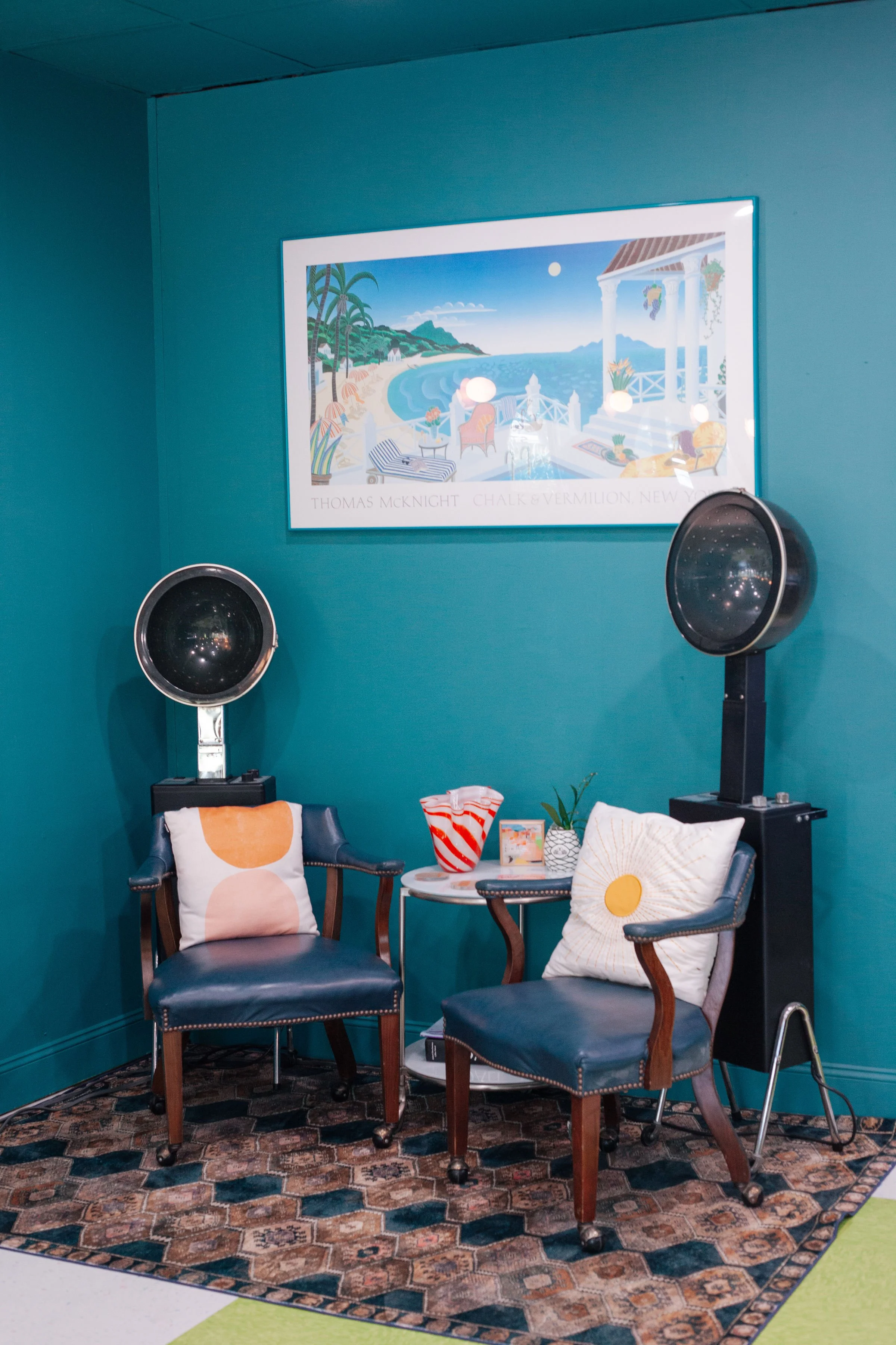 A cozy sitting area with two blue chairs, decorative pillows, a small white table with a red and white striped vase and a small framed picture, on a patterned rug. Two hair dryers are on stands behind the chairs. A framed artwork depicting a coastal 