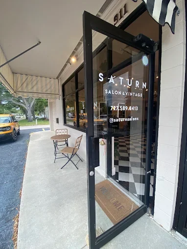 The entrance to Saturn Salon & Vintage features a glass door with the salon's name and contact info, a small outdoor seating area with two chairs, and a sidewalk outside.