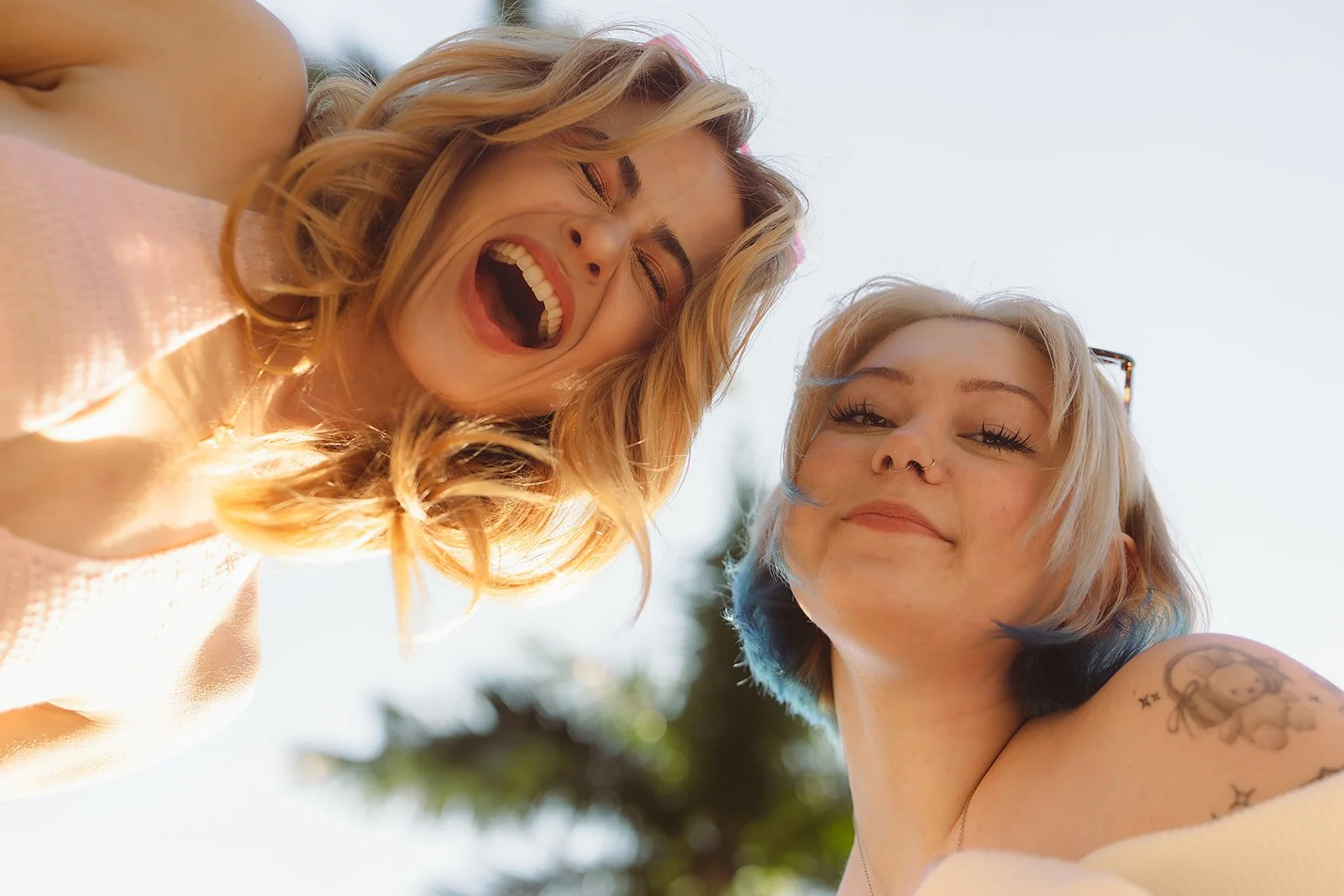 Two young women smiling and looking down at the camera with a background of trees and sky.
