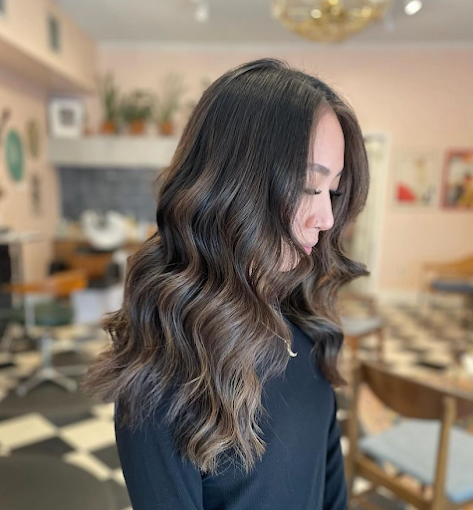 Side profile of a woman with long, wavy brown hair in a cozy, well-lit room with checkered black and white flooring, a round wooden table, and a fireplace with decorative plants in the background.