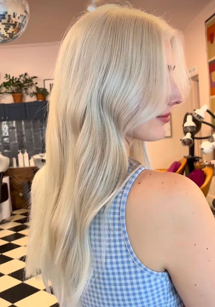 Blonde woman with long, wavy hair in a salon, wearing a blue and white checkered sleeveless top.