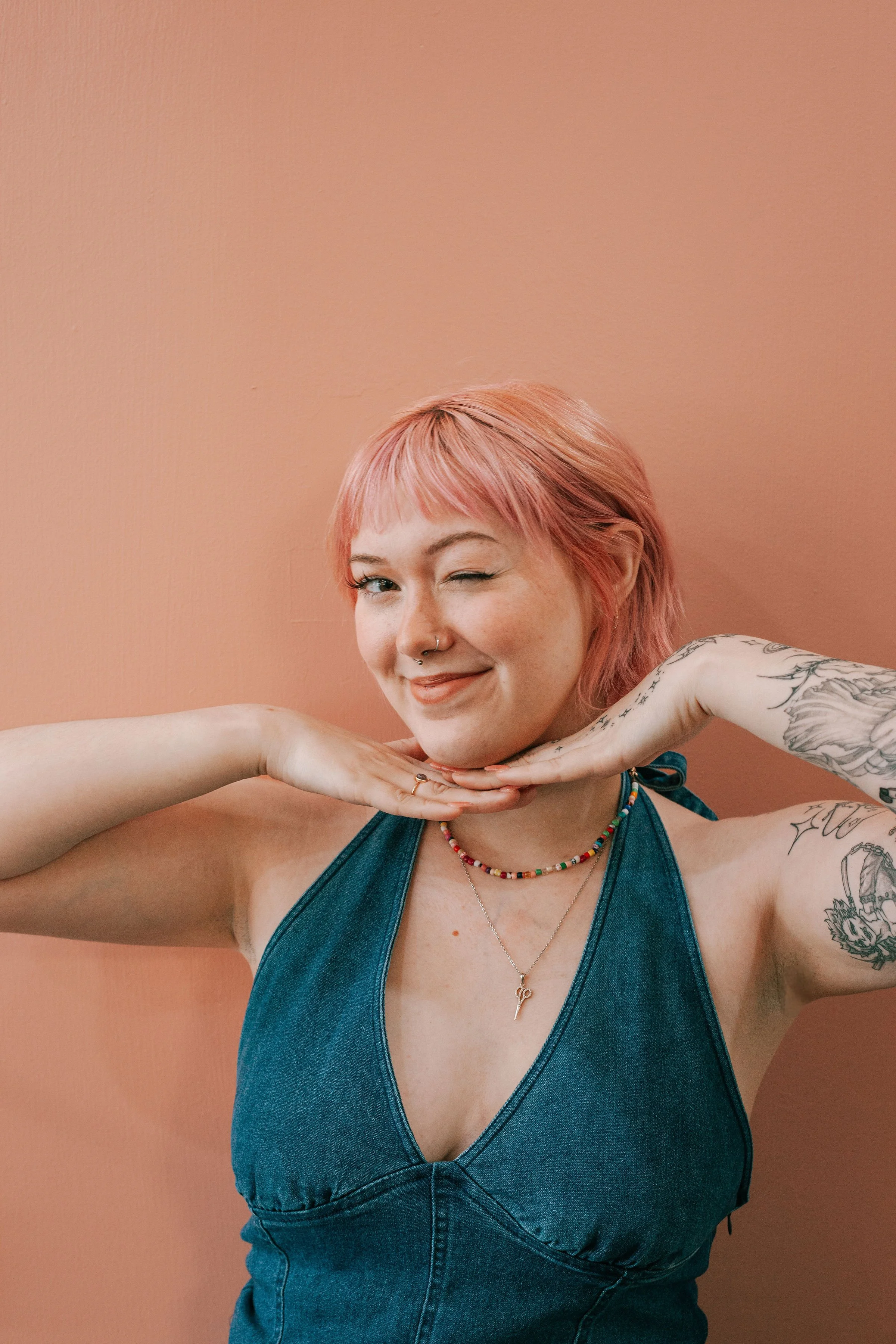 A woman with pink hair and tattoos on her arms smiling and winking, wearing a blue denim dress, colorful beaded necklaces, and a silver necklace with a lightning bolt pendant, set against a peach-colored wall.