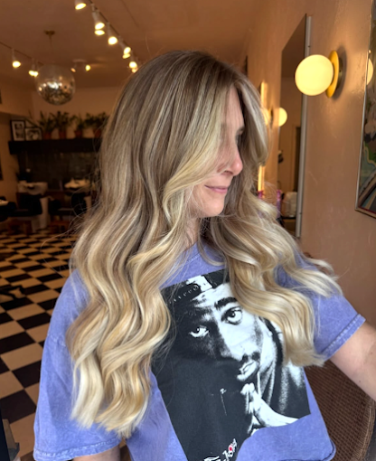 A woman with long, wavy blonde hair wearing a blue T-shirt with a black and white graphic of a woman's face, standing inside a room with warm lighting, checkered flooring, and wall-mounted lamps.