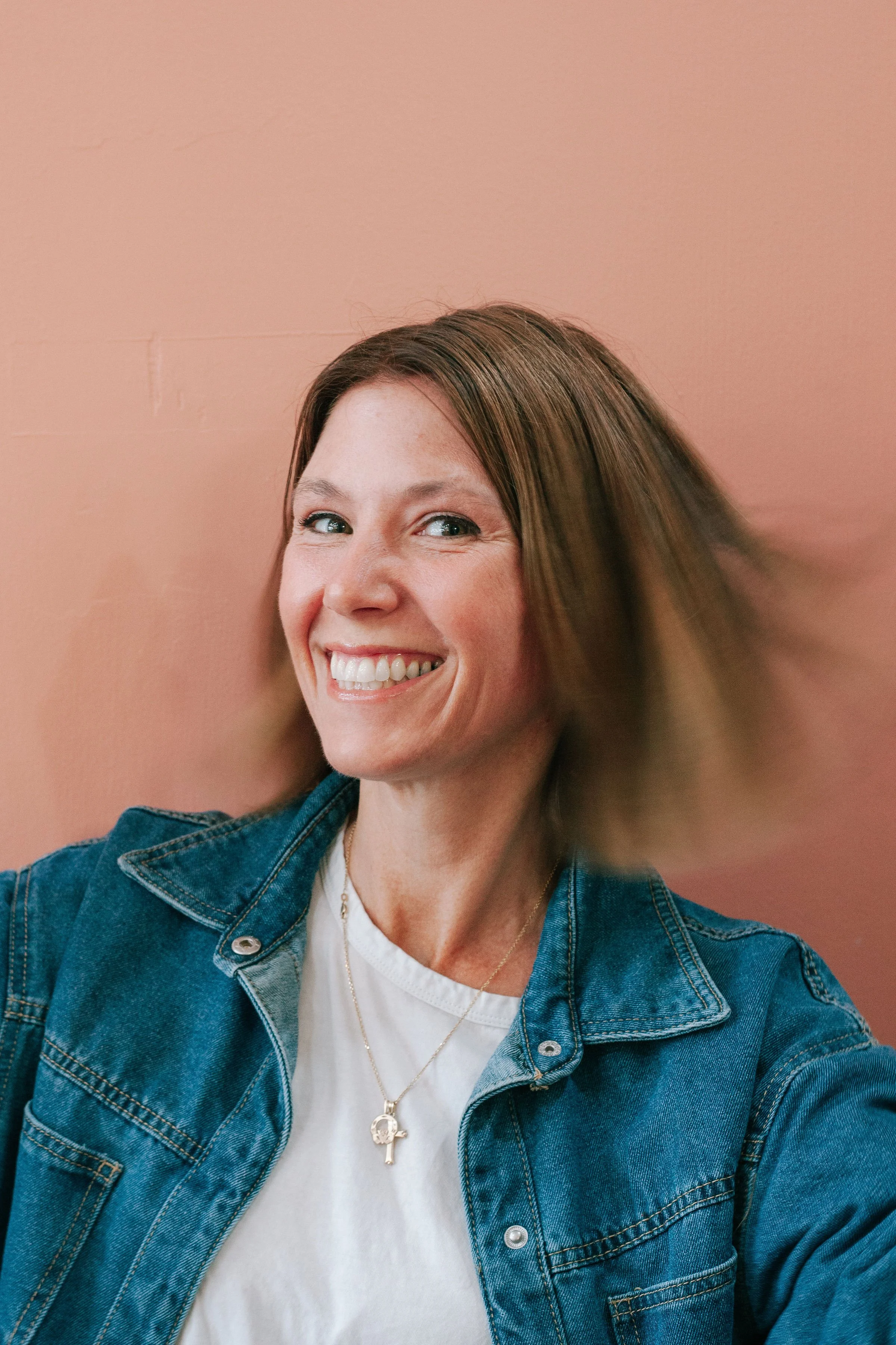 A smiling woman with brown hair in a denim jacket and white t-shirt posing against a peach-colored wall.