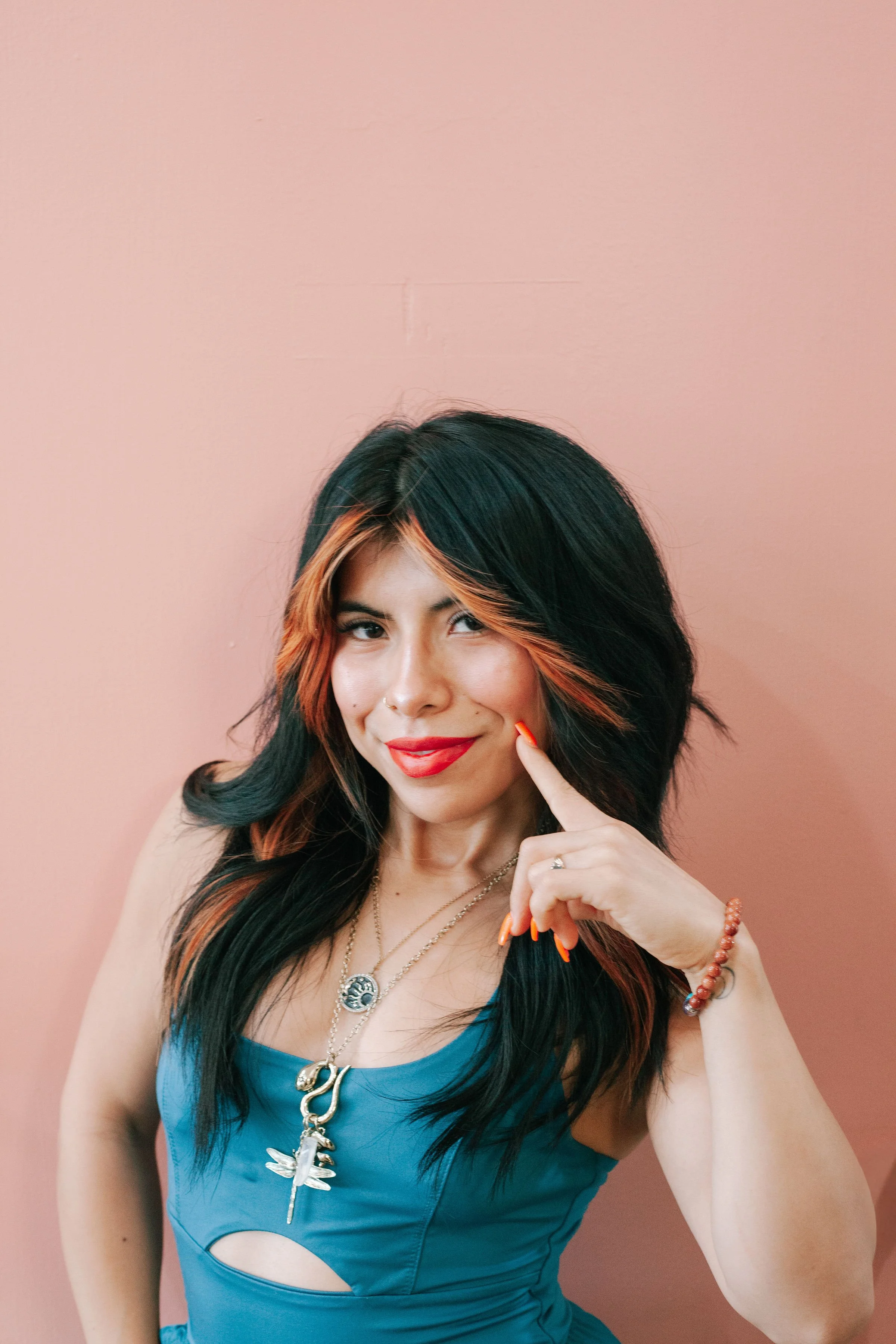 A woman with black and orange hair, red lipstick, and a blue dress posing against a pink wall, touching her face with her right hand.