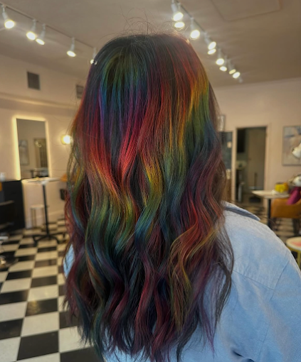 Back of a person's head showing long, wavy hair colored with rainbow hues, in an indoor setting with checkered floor and modern decor.