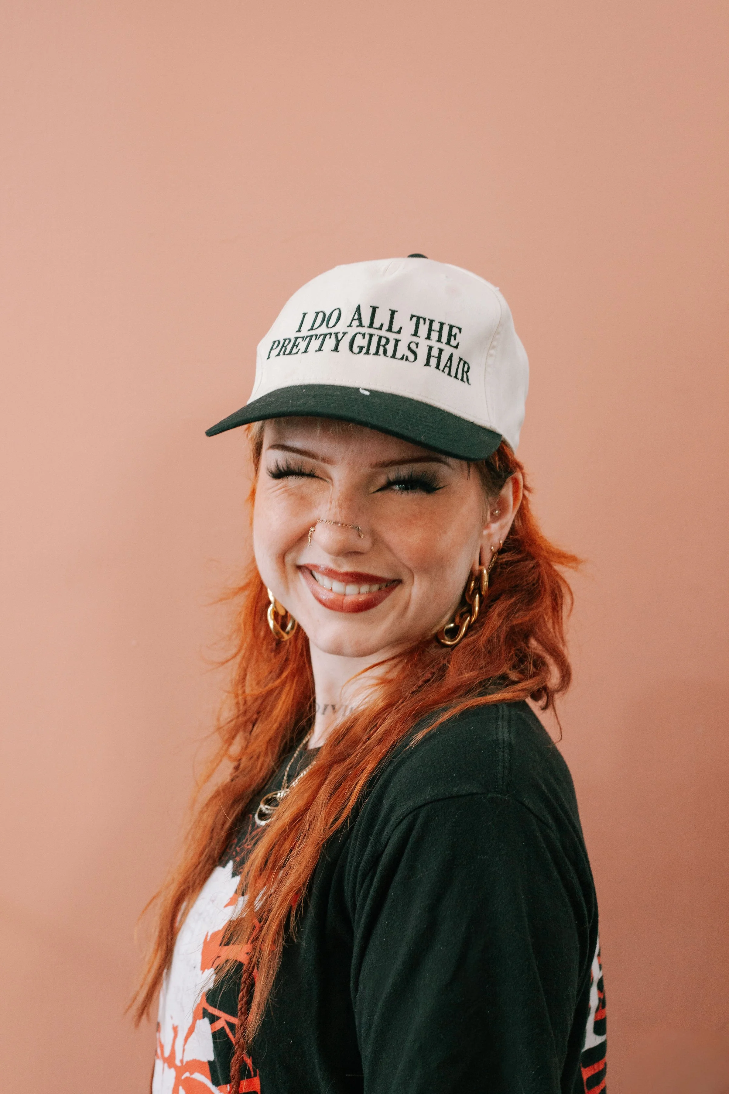 A woman with red hair, wearing large gold hoop earrings, a white baseball cap with green text, and a black shirt, smiling against a pink wall background.