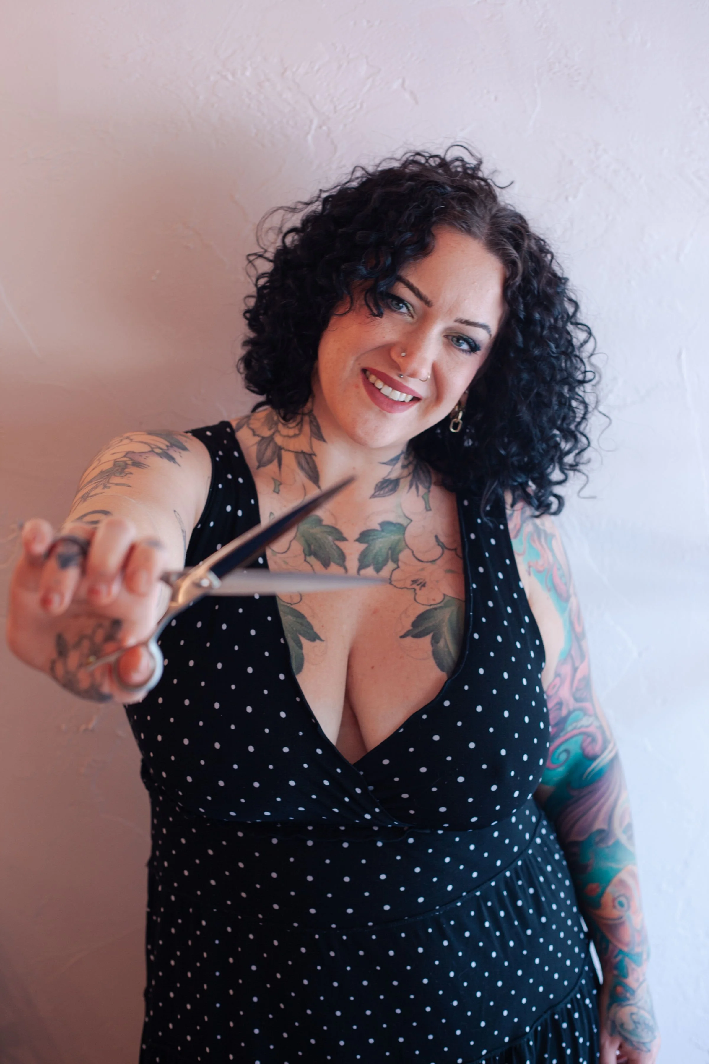 A woman with curly black hair taking a selfie, wearing a black polka-dot dress with a plunging neckline, and has tattoos on her chest, arms, and shoulders, standing against a plain white wall.
