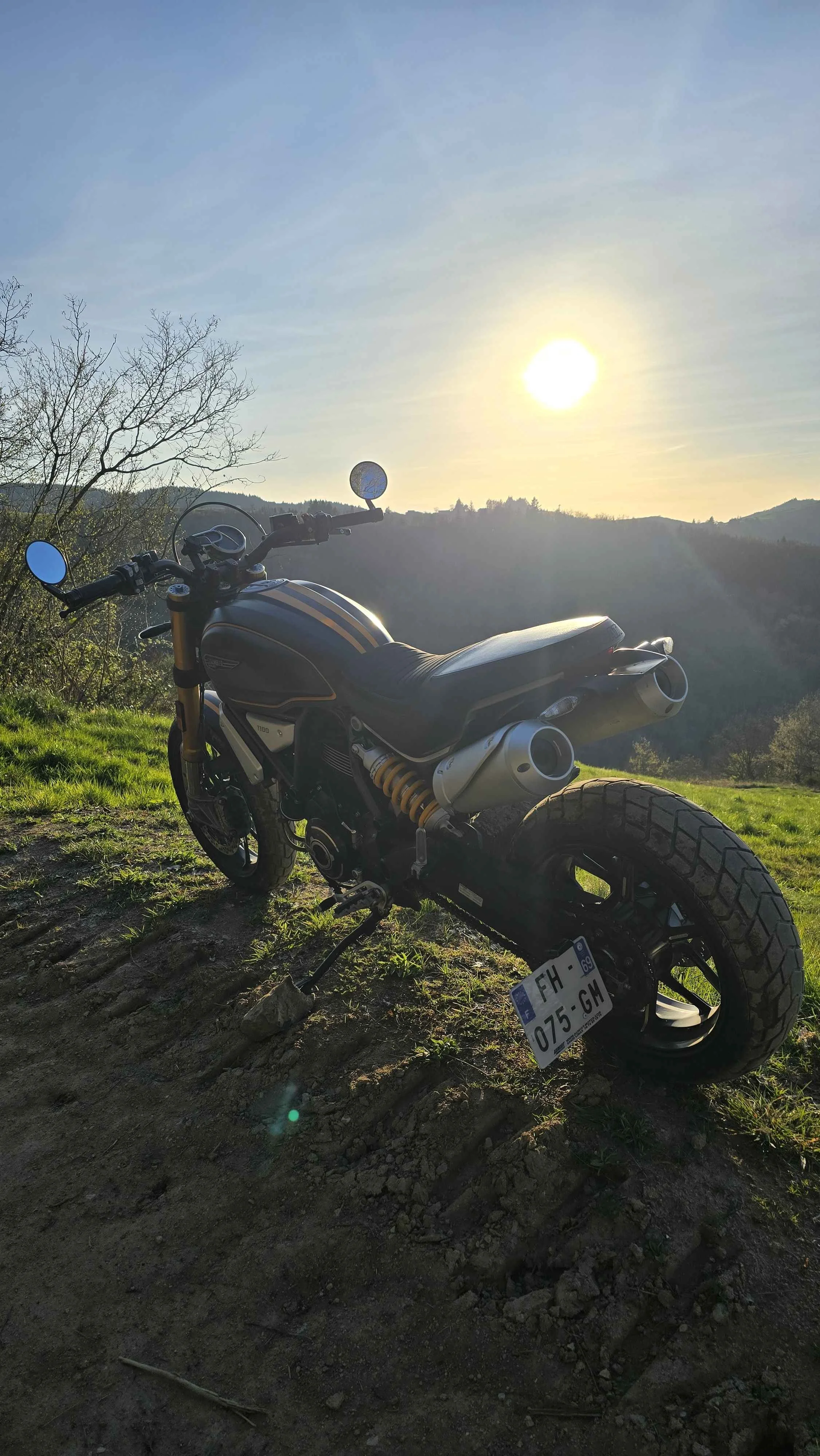 Une moto stationnée sur un terrain en terre avec des montagnes en arrière-plan, sous un ciel lumineux avec le soleil brillant haut dans le ciel.