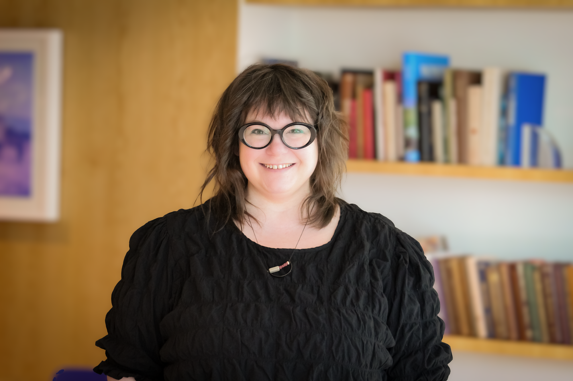 A headshot of TRNSLATE Studio's founder, Allison Stratton. She has thick-rimmed black glasses and a black blouse with a minimalist necklace. Her hair and eyes are brown.