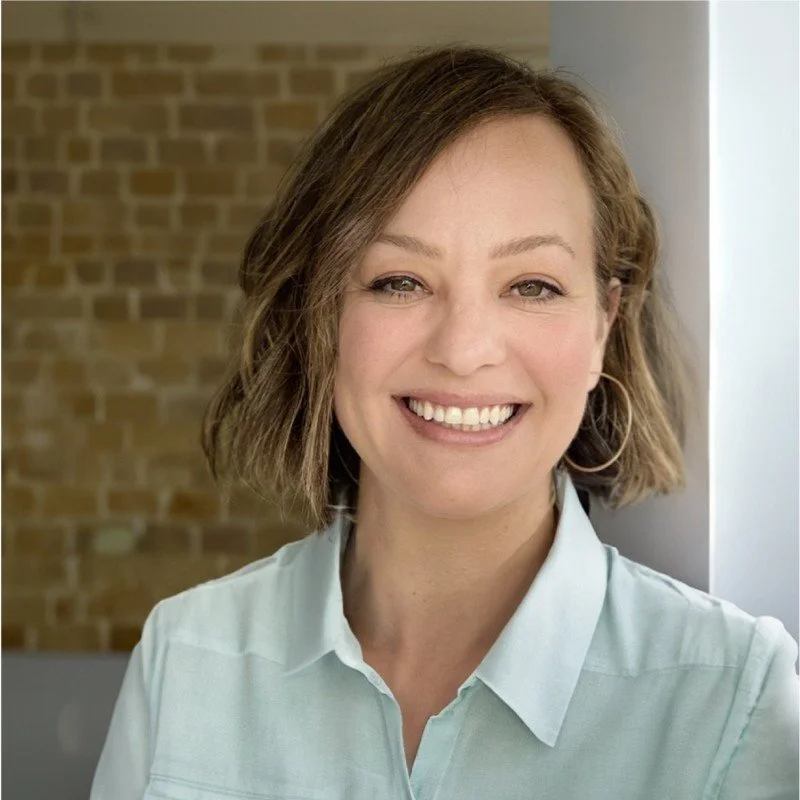 Close-up of a woman smiling, wearing a light-colored blouse, with short, wavy brown hair, in front of a brick wall.