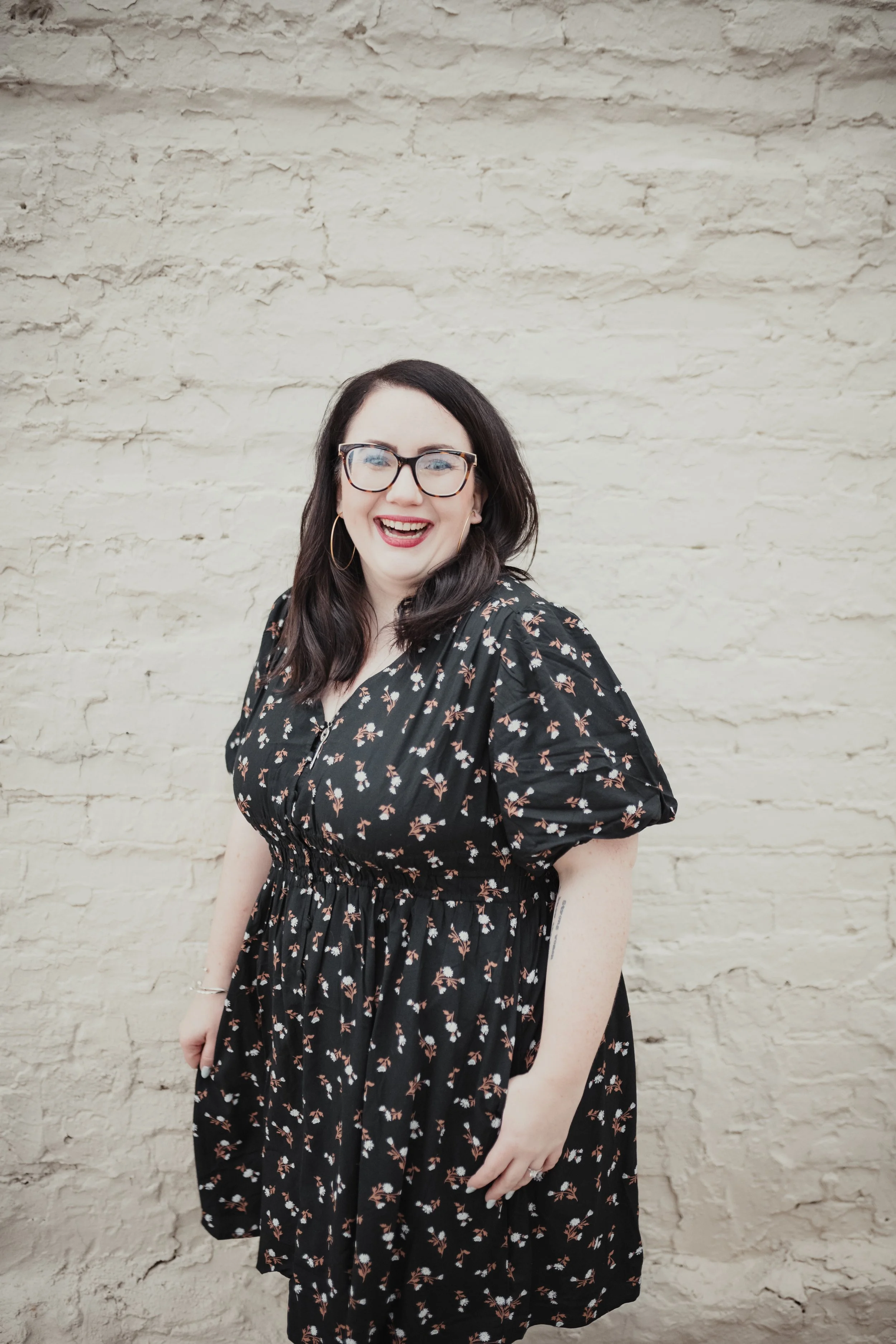 A woman with dark hair, glasses, and hoop earrings smiling while wearing a black floral dress, standing in front of a light-colored brick wall.
