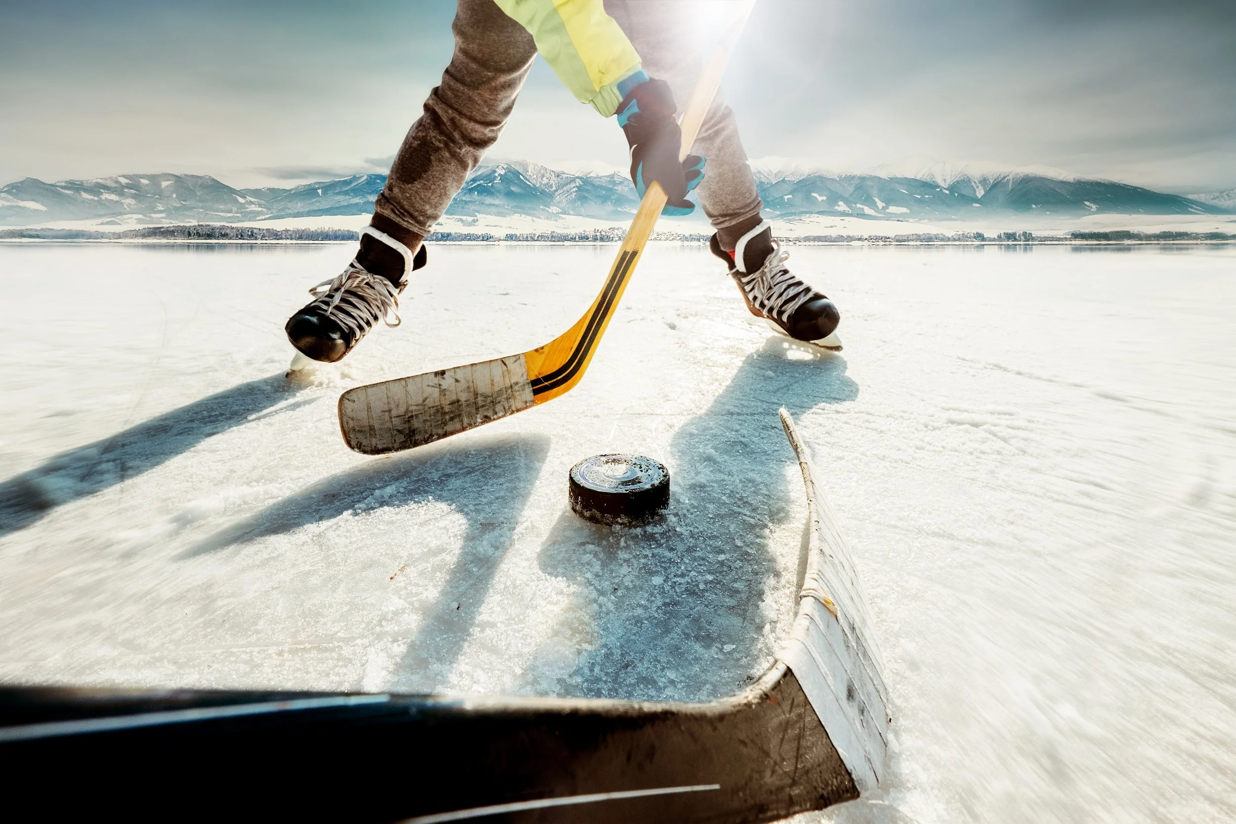Person skating on ice outdoors with mountains in the background, using a hockey stick and puck on the ice.