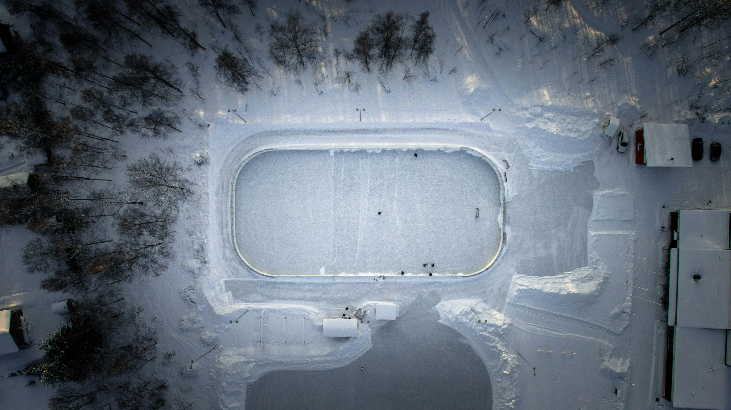 A snow-covered outdoor ice skating rink surrounded by leafless trees and parking lot, viewed from above.