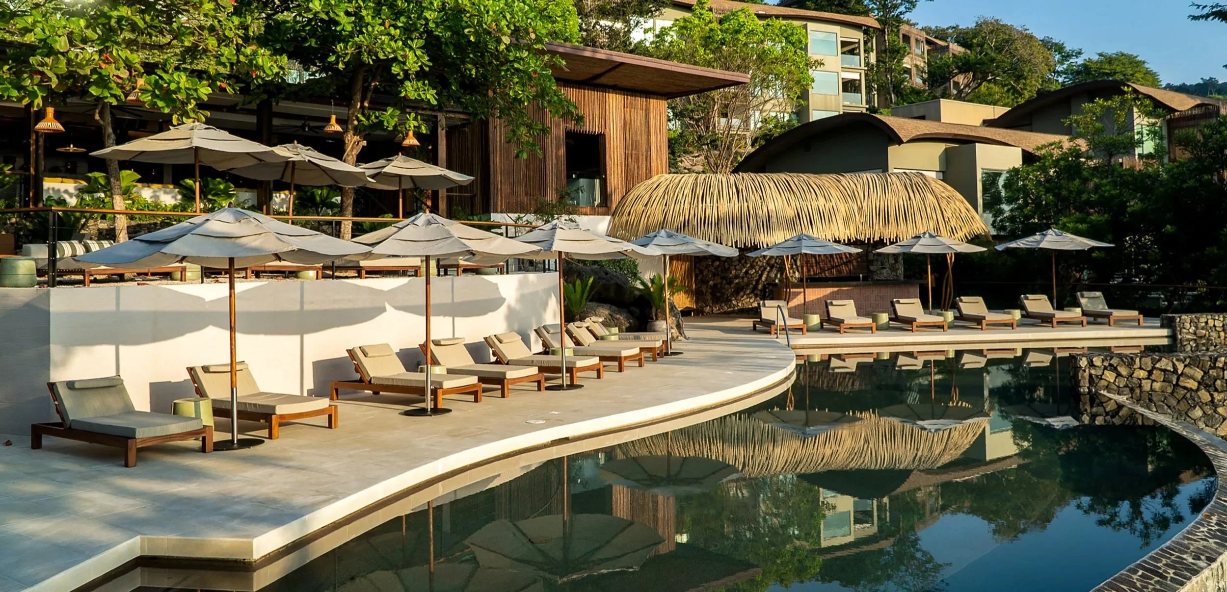 Luxury resort pool area with lounge chairs, umbrellas, lush green trees, and modern buildings in the background.