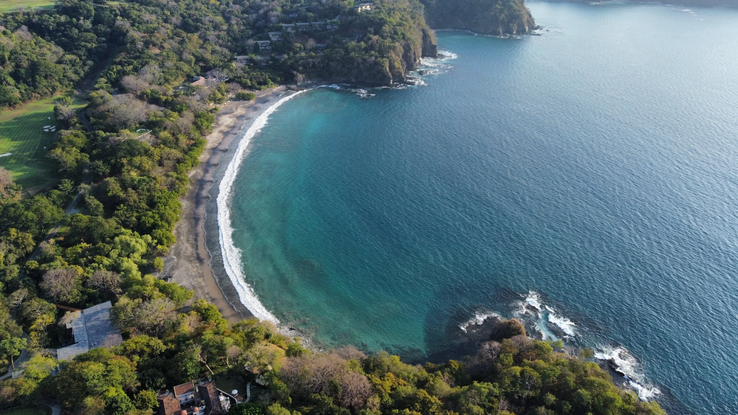 Aerial view of a coastline with a beach, clear blue water, lush green trees, and rocky cliffs.