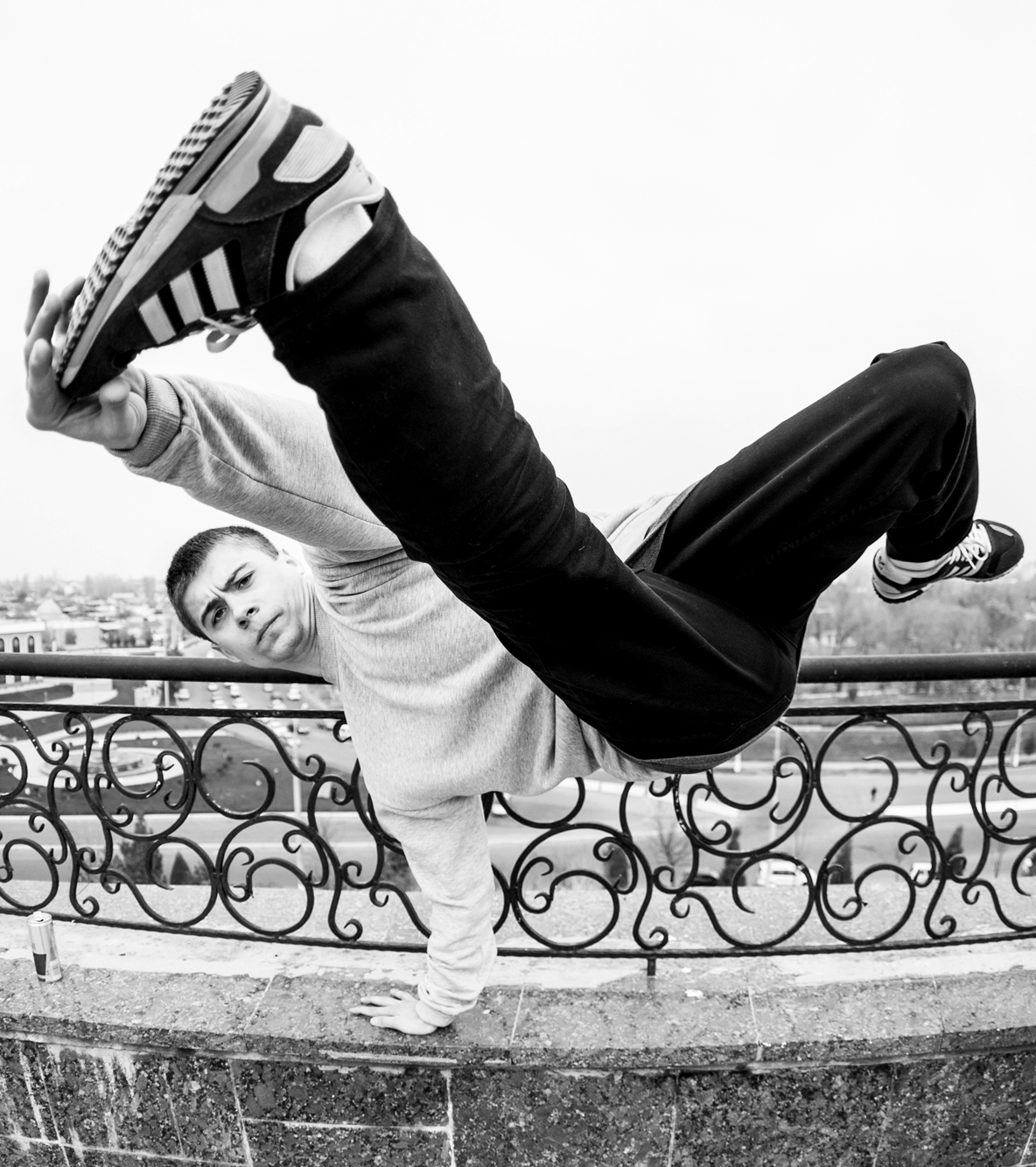 A young man performing a one-handed handstand on a park ledge, with a cityscape background in black and white.