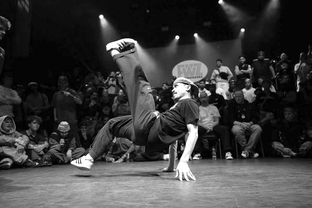 A breakdancer in a black shirt and pants performs a move on the dance floor while an audience watches in the background at a dance competition.