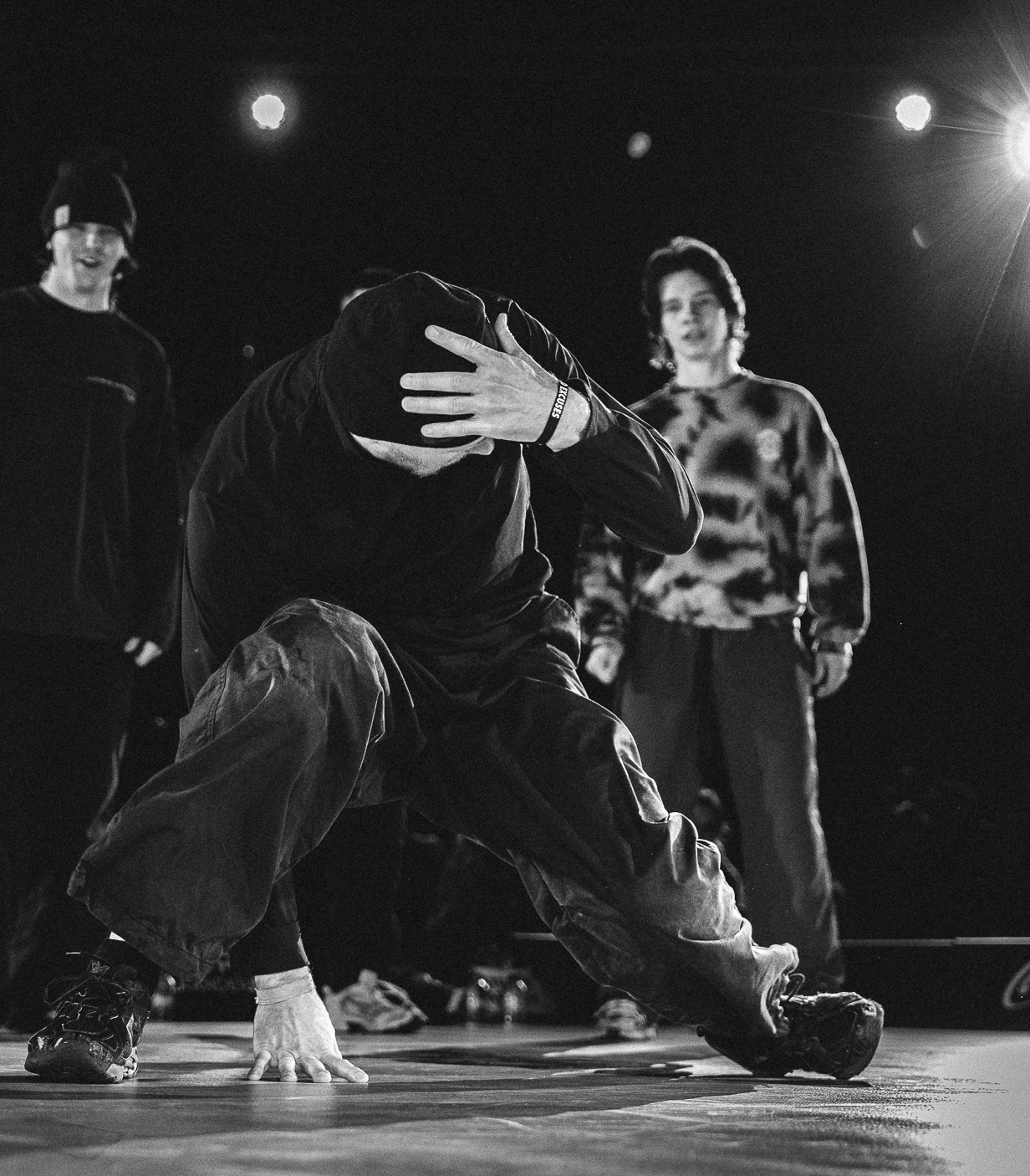 A breakdancer is crouched on one hand and knee, holding his head with one hand, in a dark space with bright lights. Two other performers or audience members stand behind him, watching.