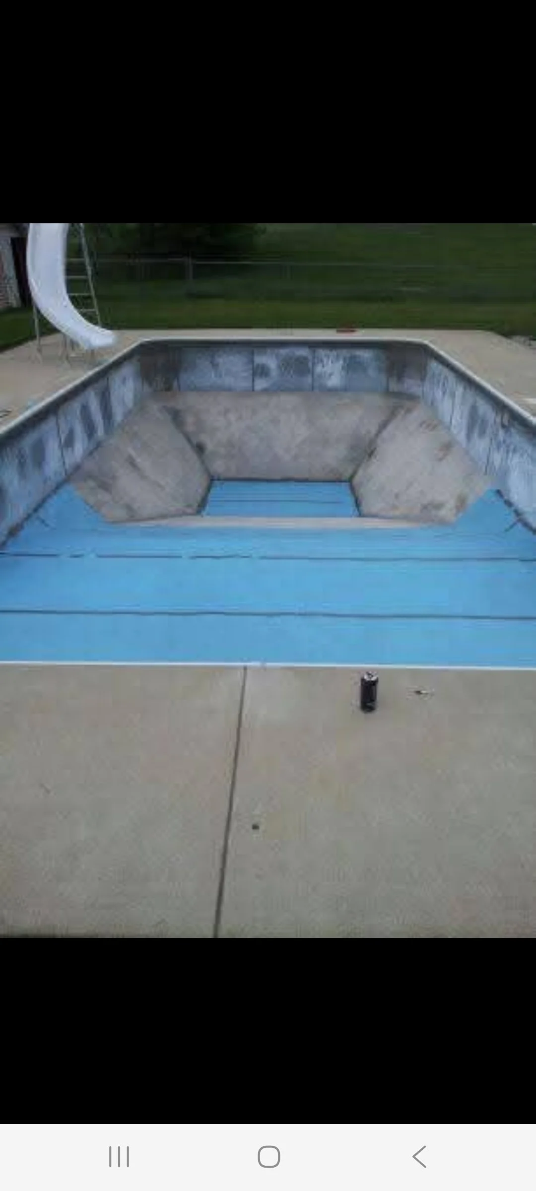 Empty backyard swimming pool with a slide, surrounded by a concrete deck, with a grassy yard and trees in the background.