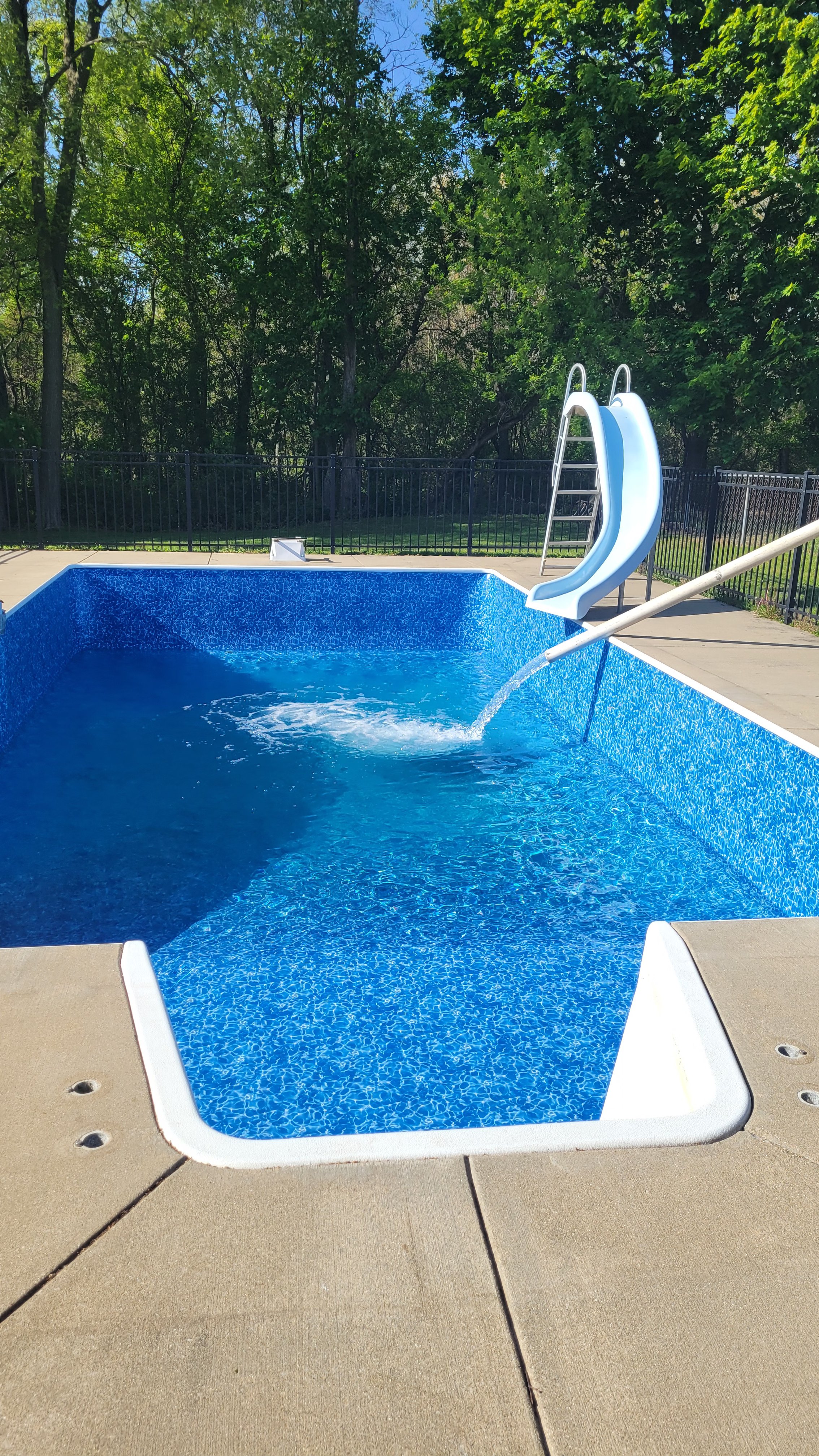 Empty swimming pool with a water slide and a poolside lounge chair, surrounded by a fence and trees.