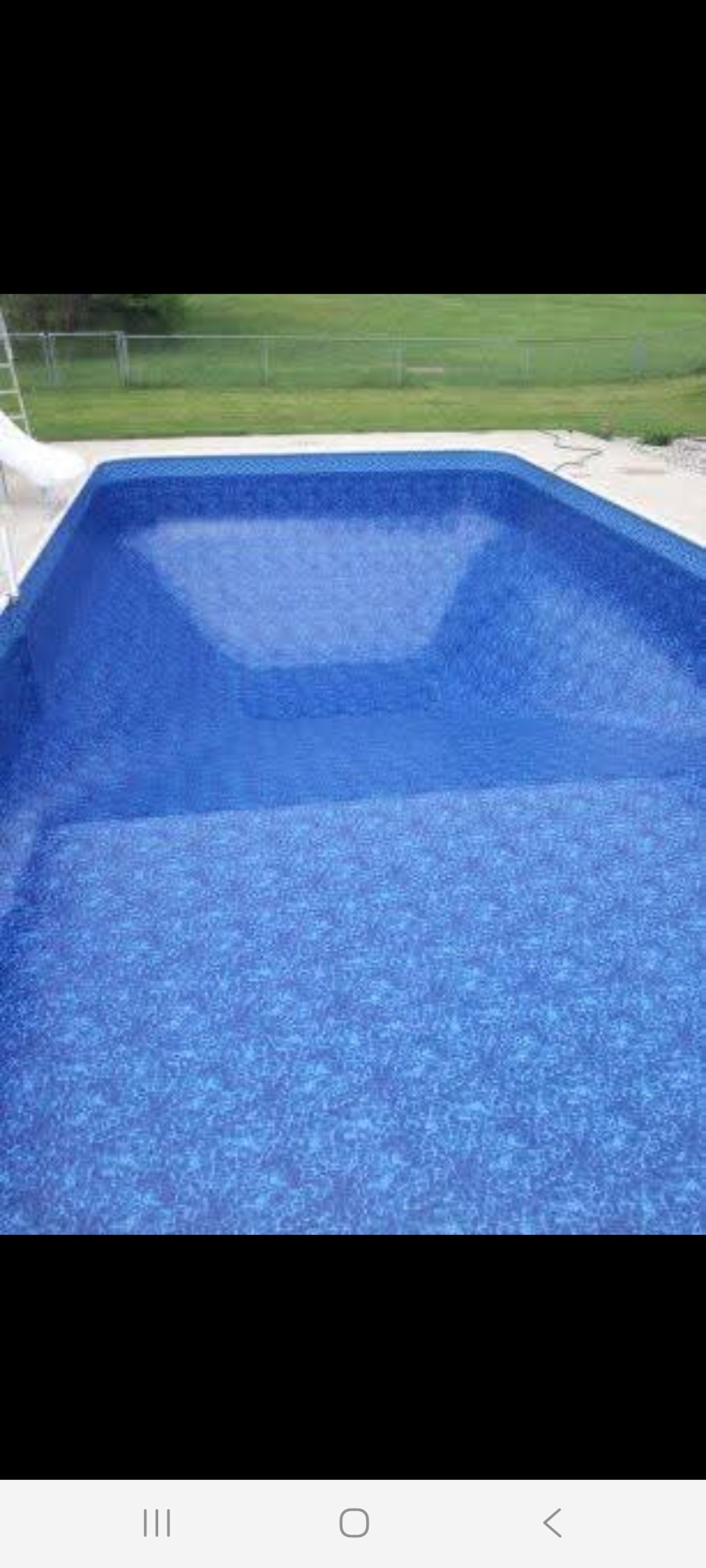 A rectangular swimming pool with a vibrant blue interior, situated in a backyard with green grass and a metal fence in the background.
