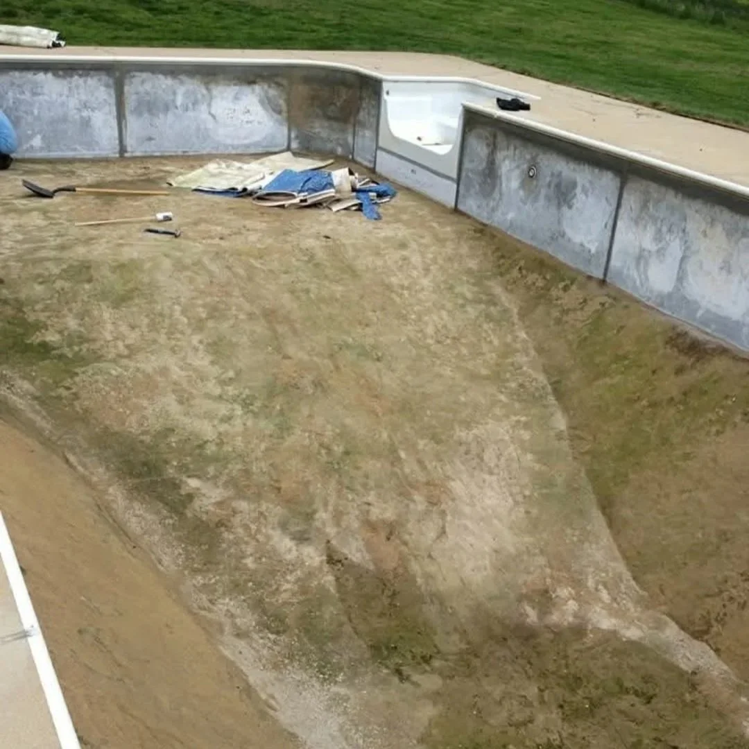 An empty skateboard pool or mini ramp under construction, with dirt and some construction tools and materials inside, and surrounded by grass.