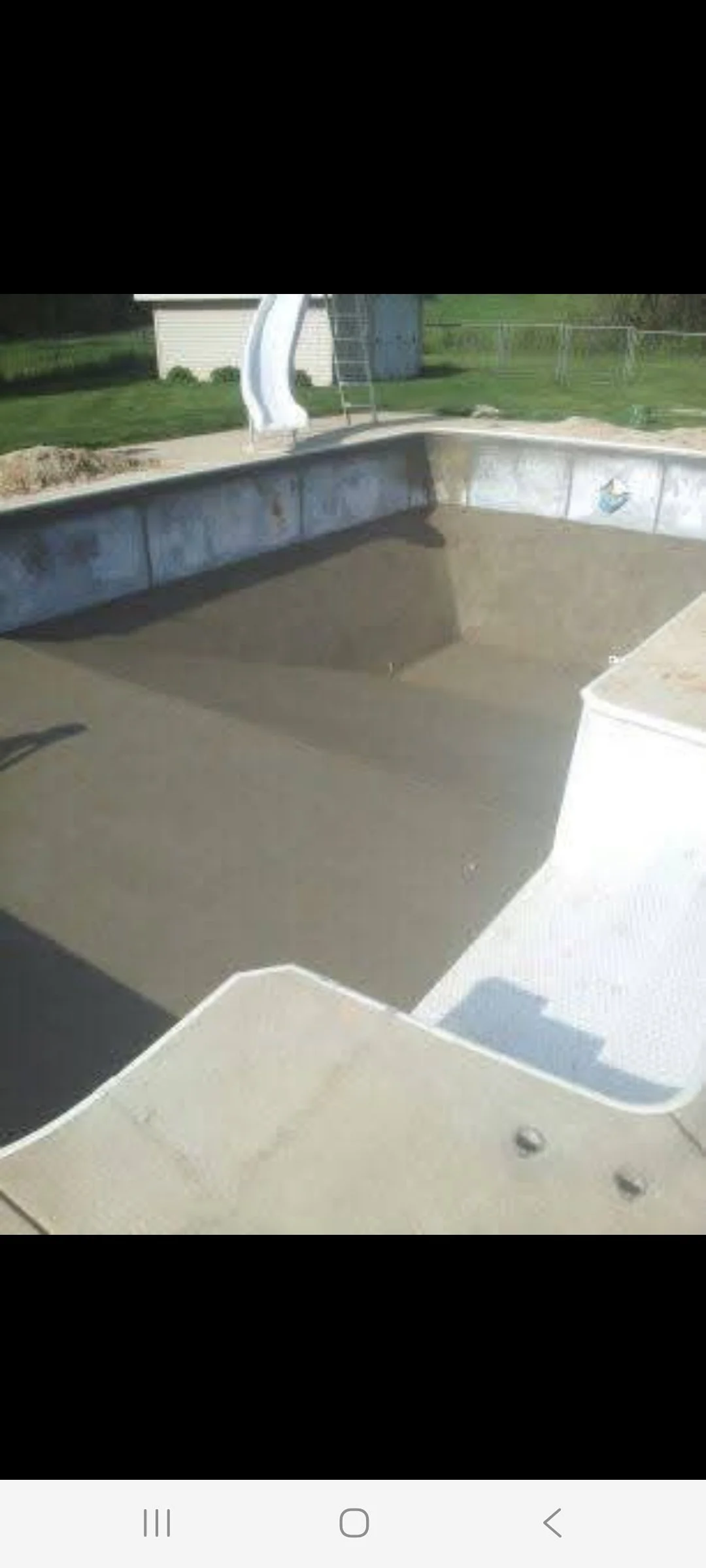 Empty in-ground swimming pool with a water slide in a backyard, surrounded by grass, a small shed, and a few outdoor structures.