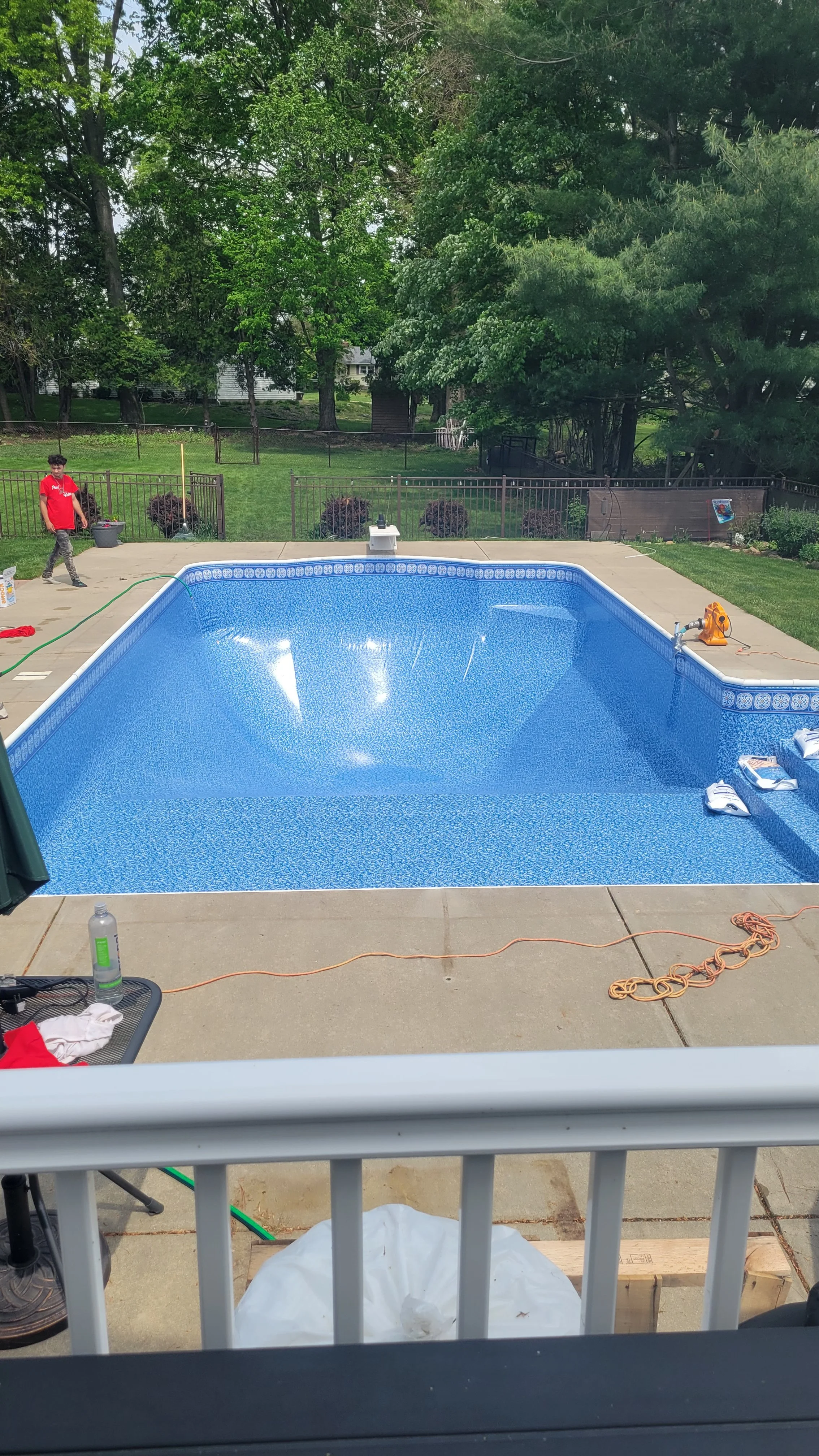 Backyard with a rectangular in-ground swimming pool, partially filled with water, on a concrete patio. A person in a red shirt is walking on the left side, and various pool tools and supplies are scattered around. In the background is a grassy yard with trees and a fence.