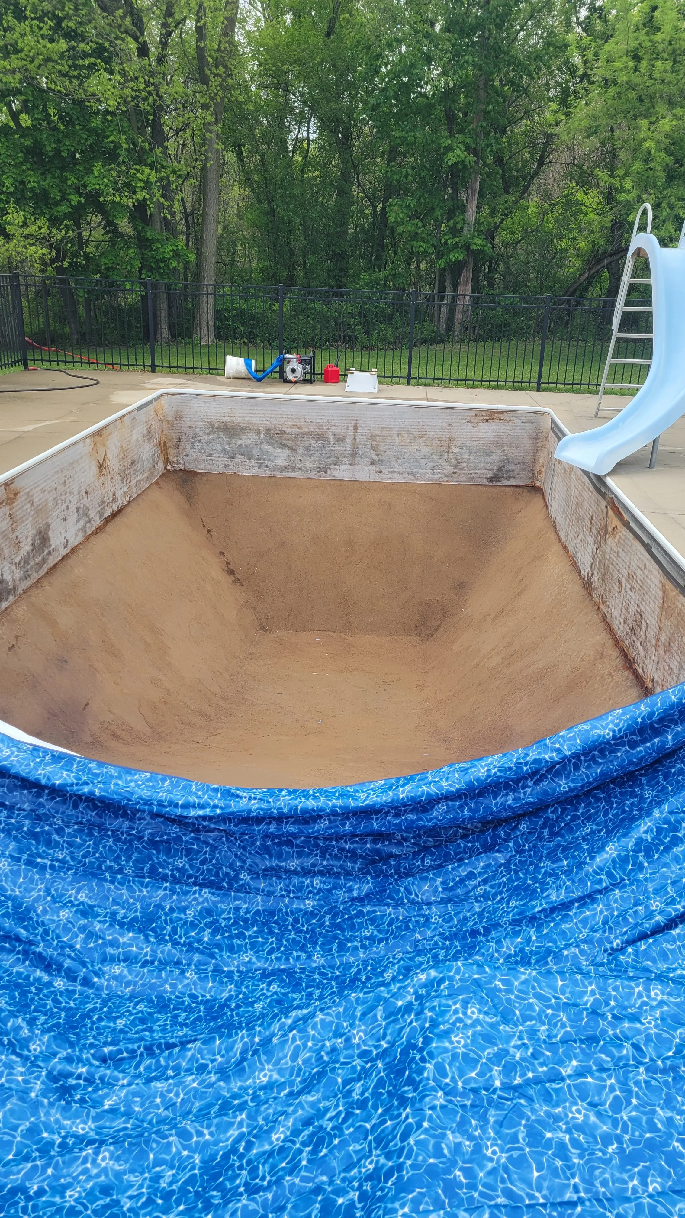 Empty swimming pool with a beige interior, surrounded by a blue pool cover, on a wooden deck. In the background, there is a slide, a fence, green trees, and pool cleaning equipment.