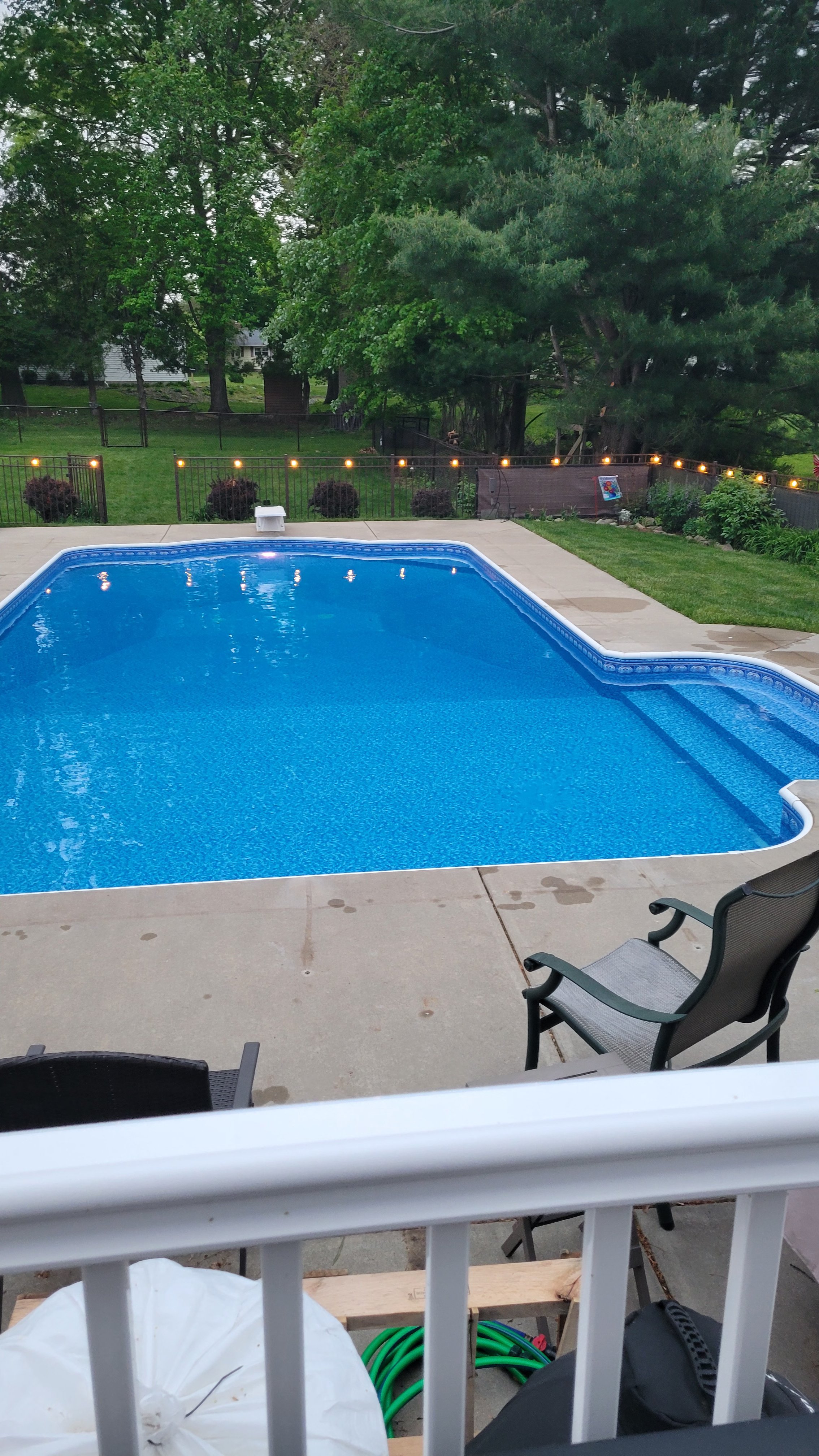 View of a backyard with a swimming pool, surrounded by a concrete patio and surrounded by green trees and bushes. There is a string of lights along the fence, and outdoor furniture including a chair in the foreground and a lounge chair on the patio.