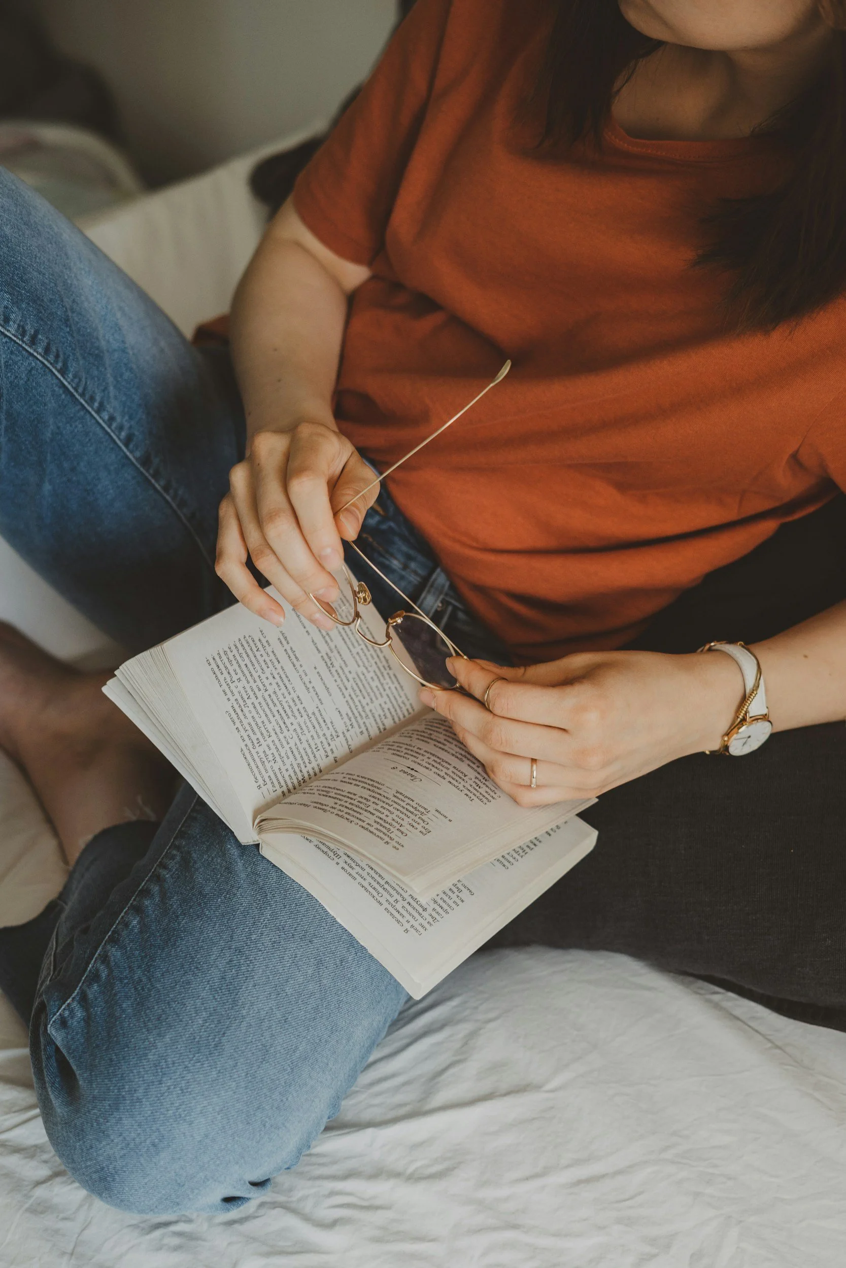 A person wearing a rust-colored t-shirt and blue jeans sitting on a bed, holding eyeglasses and a book or magazine.