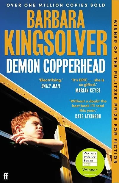 The cover of the book 'Demon Copperhead' by Barbara Kingsolver features a young boy with red hair, resting his arm on a school bus window, looking thoughtfully into the distance. The cover includes quotes praising the book and highlights awards, with blue and yellow color themes.